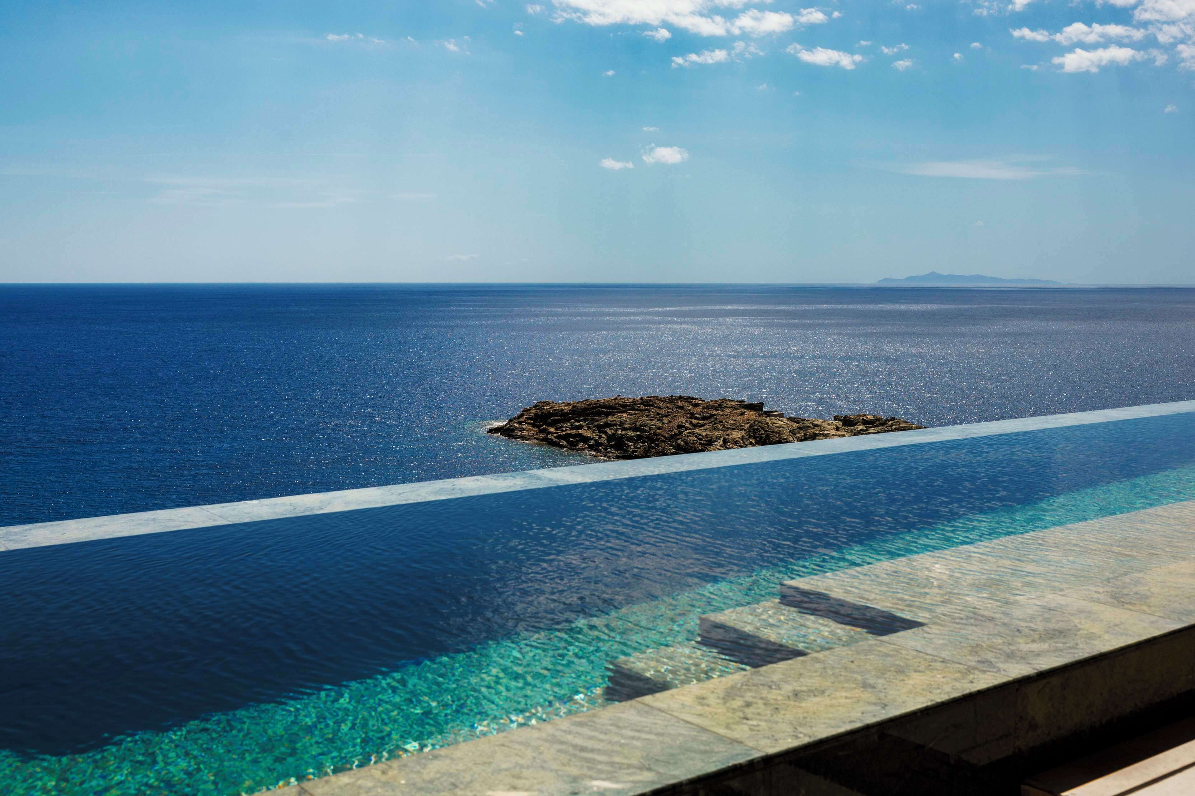 A pool with clear water, a rocky island, and a distant mountain under a blue sky.