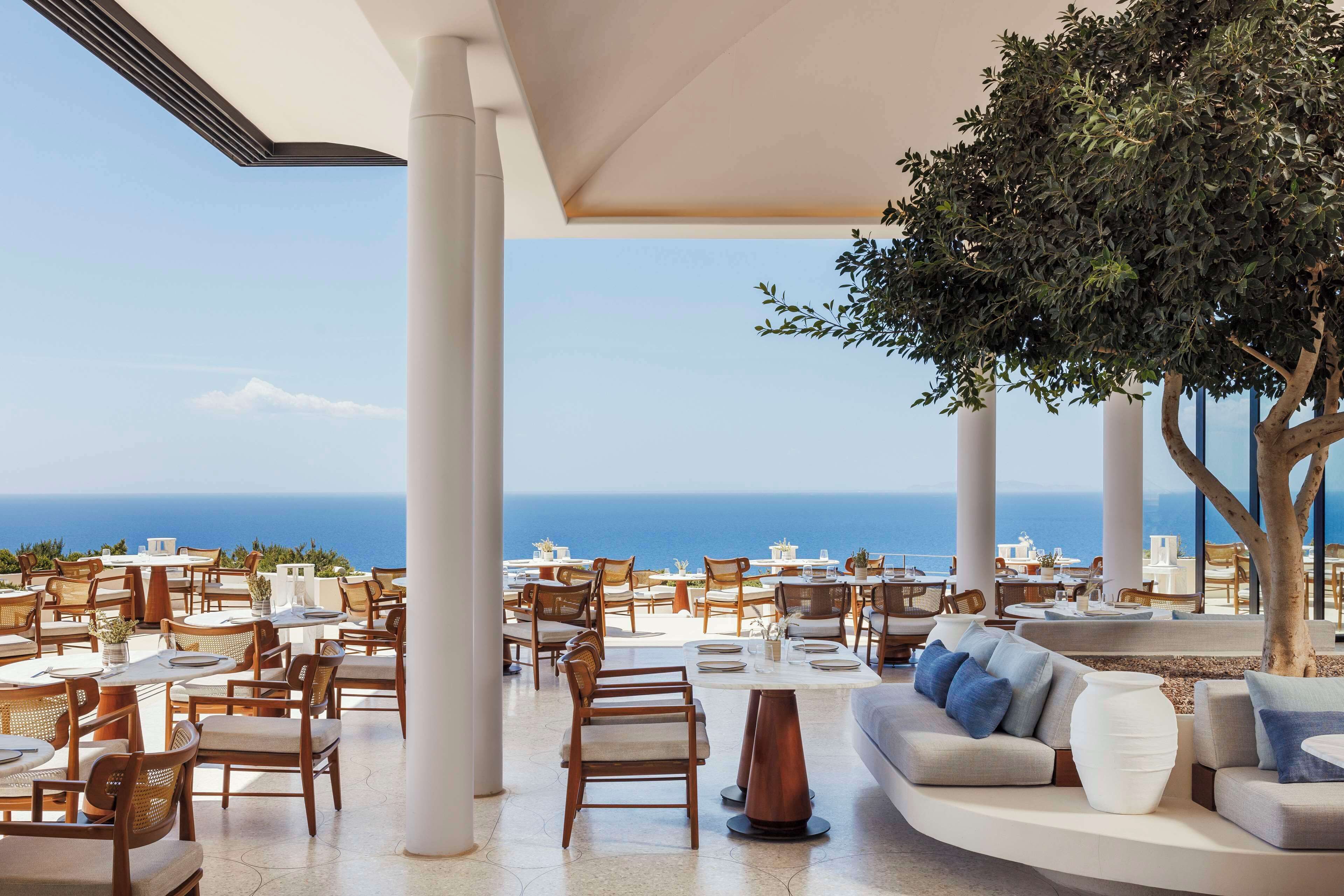 A rooftop dining area with tables and chairs, a large tree, and a view of the ocean.