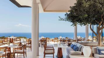 A rooftop dining area with tables and chairs, a large tree, and a view of the ocean.