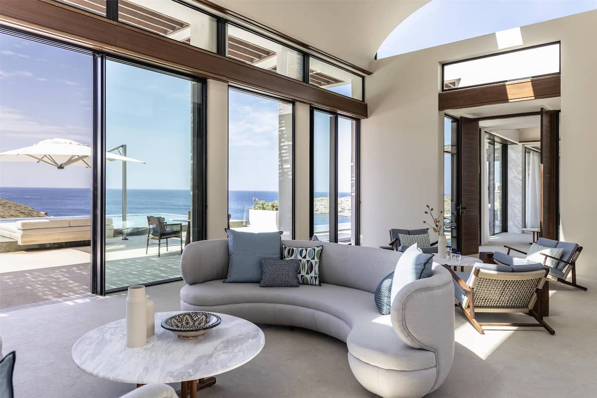 A modern living room with a curved sofa, wicker chairs, a marble table, and large windows overlooking the ocean.