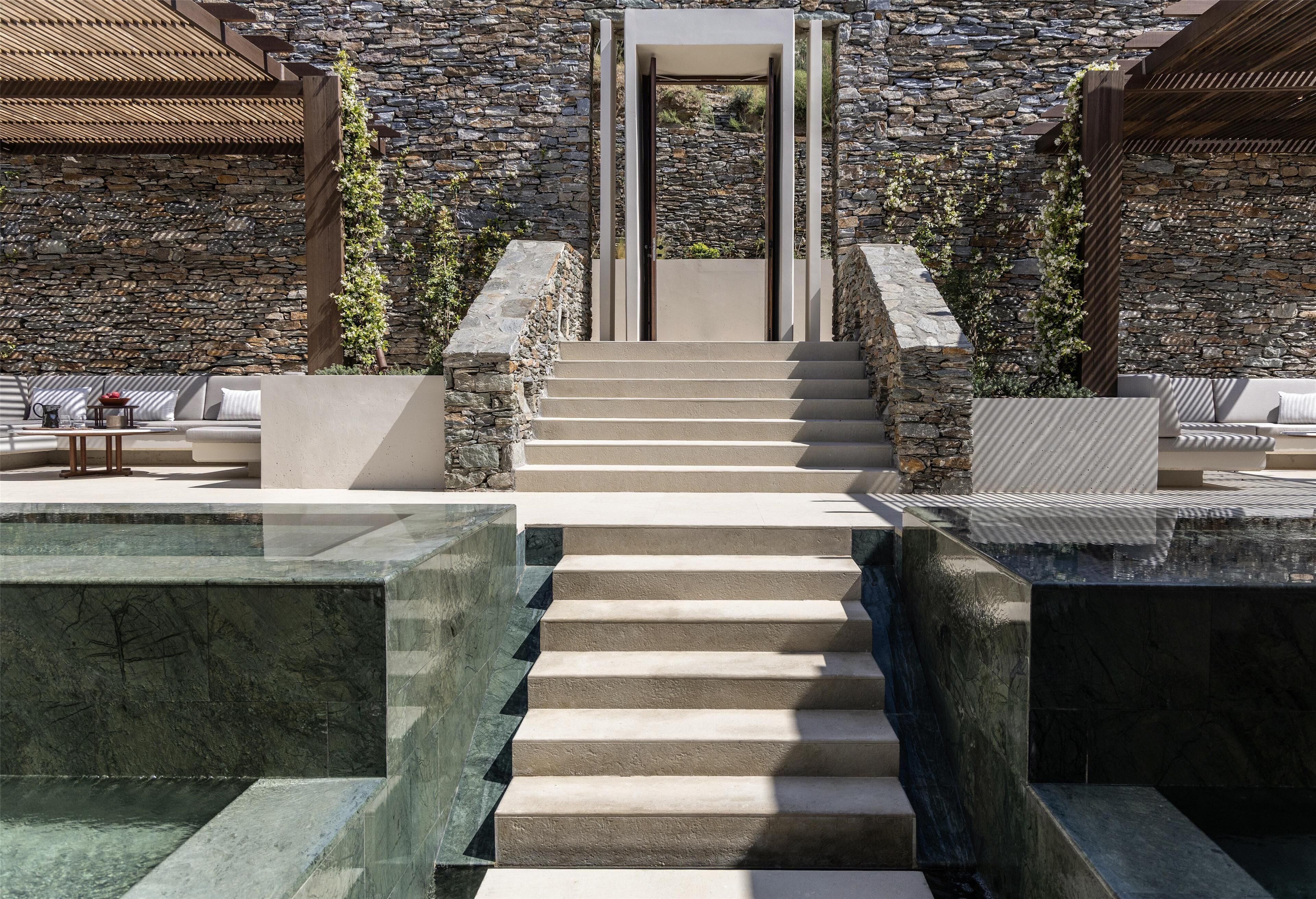 A stone-clad building with a staircase leading to a door, outdoor seating, and a water feature.