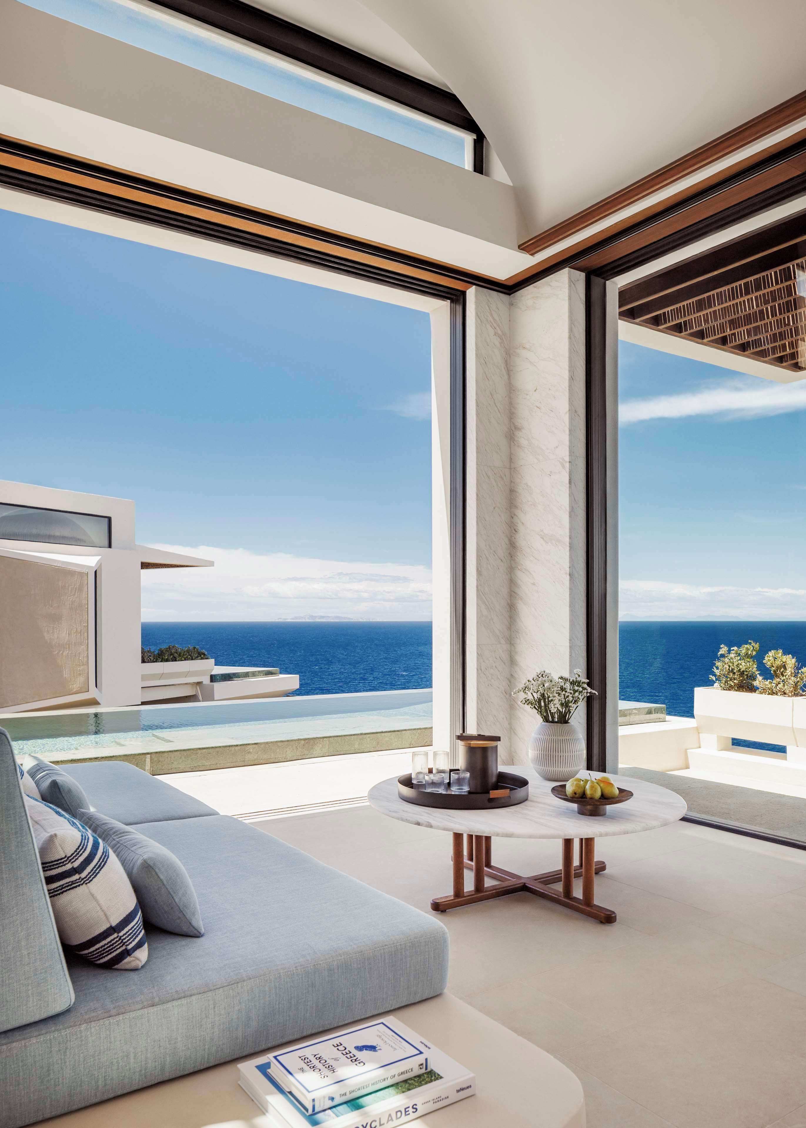 A modern living room with a large window overlooking the sea, a round table with a vase and glasses, and a sofa with cushions.