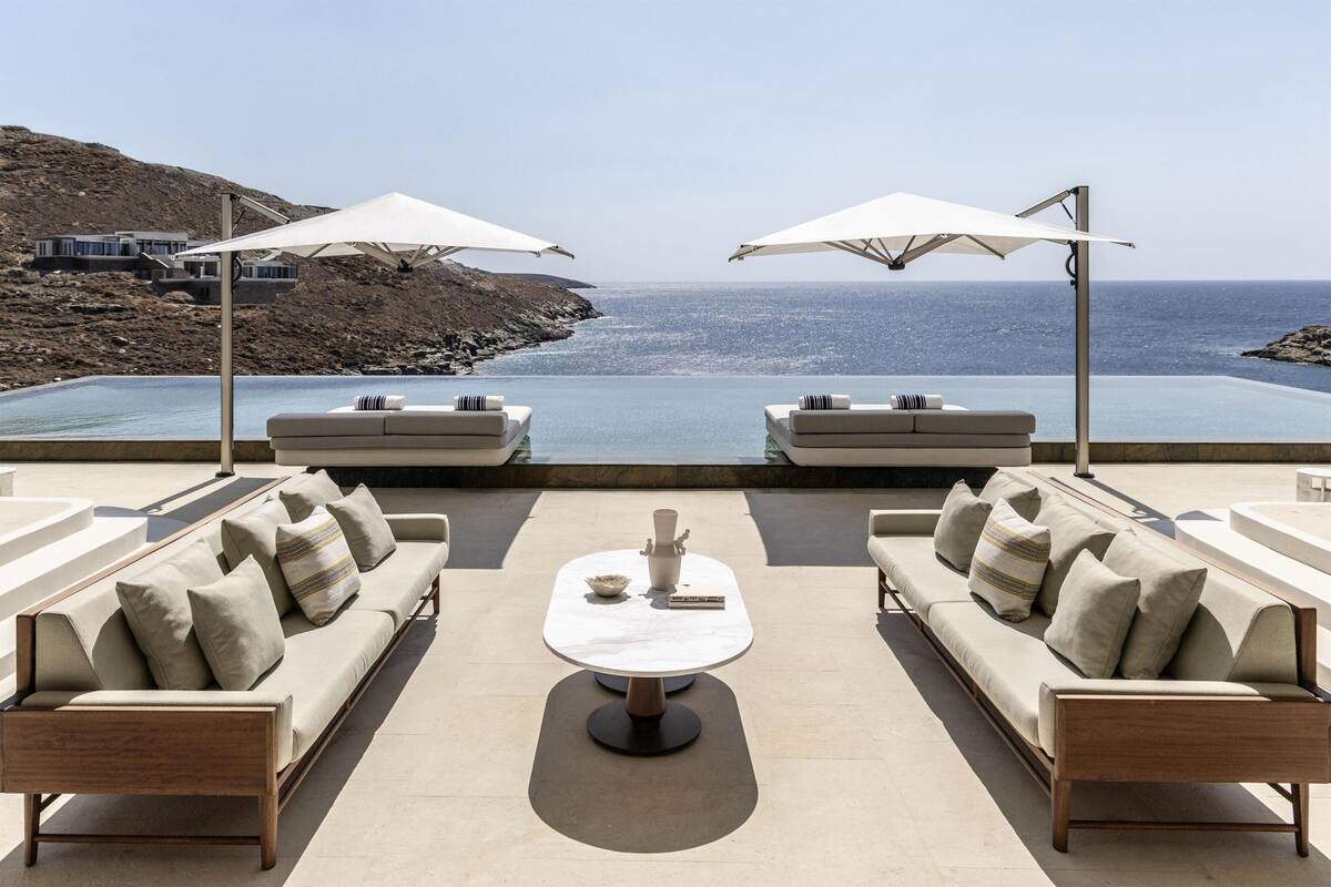 A modern outdoor seating area with a view of the sea, featuring sofas, a table, and umbrellas.