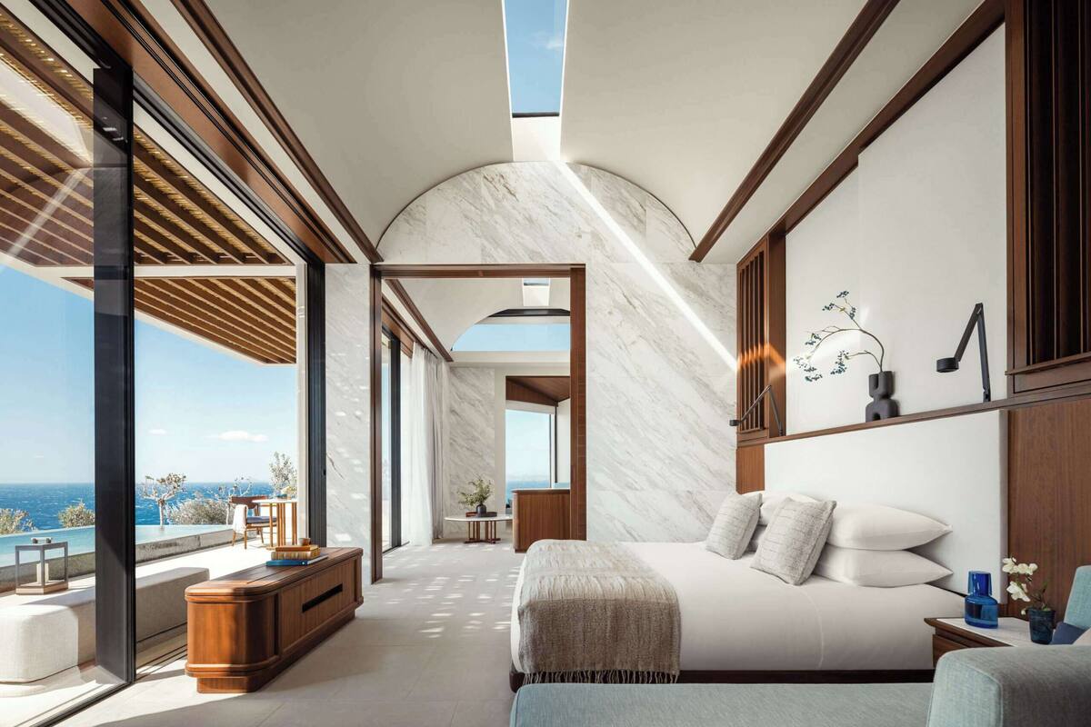 A bedroom with a large bed, a wooden bench, a small table, and a view of the ocean.