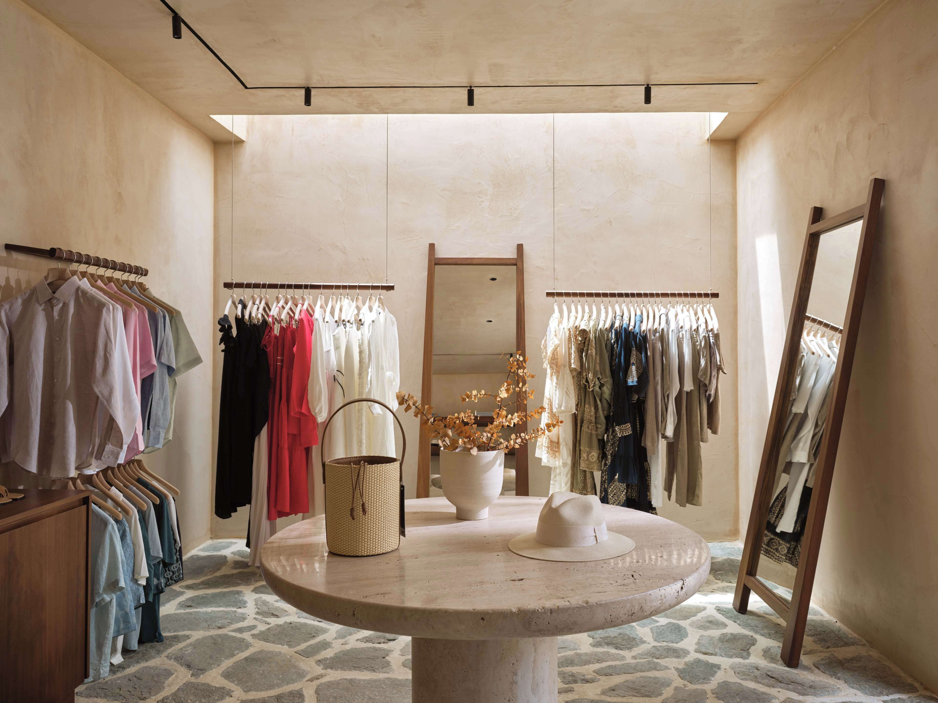 Une boutique avec des portants à vêtements, une table ronde sur laquelle est posé un chapeau, et un miroir.