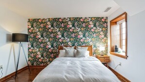 Een slaapkamer met bloemen behang, een bed met witte lakens, een nachtkastje met een lamp, en een raam met uitzicht.