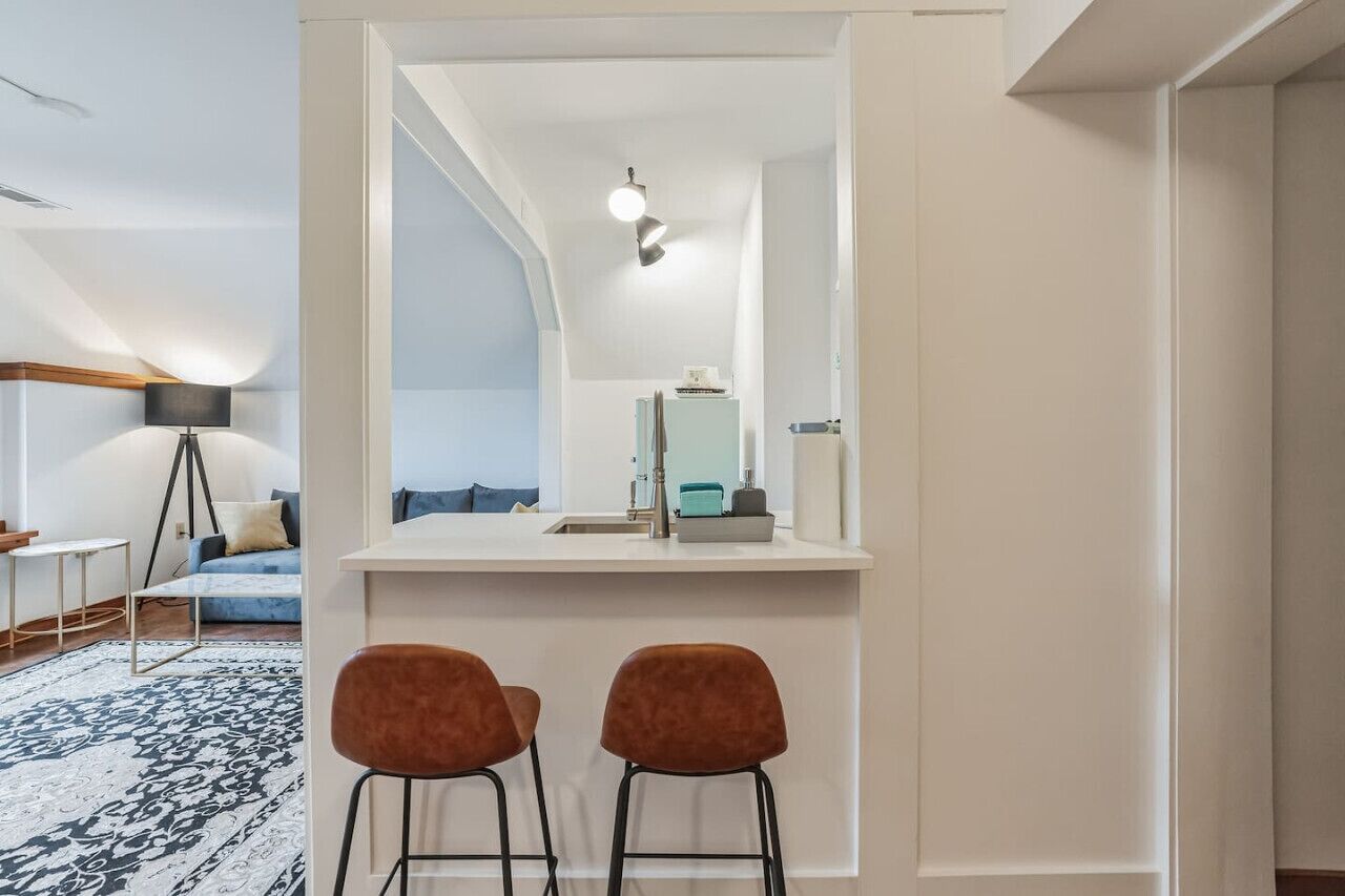 A modern kitchen with a white countertop, two bar stools, and a hanging light fixture.