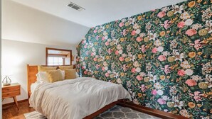 A bedroom with a floral wallpaper, a wooden bed frame, and a nightstand with a lamp. - Thornton Studio w/ Kitchenette (Thornton)