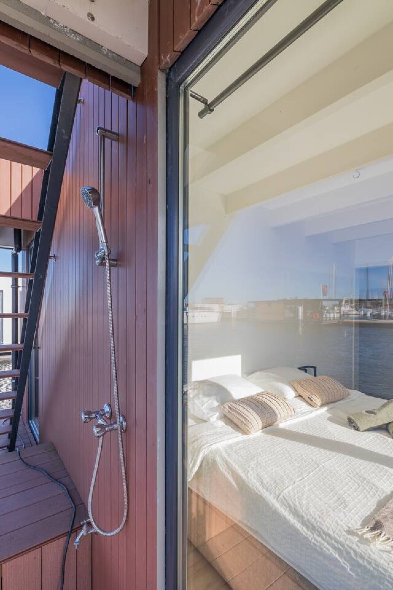 Un balcón con suelo de madera, una escalera metálica y una puerta de cristal que da a un dormitorio con cama y almohadas.