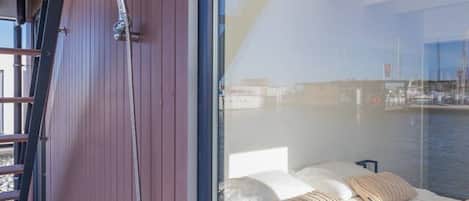 A balcony with a wooden floor, a metal ladder, and a glass door leading to a bedroom with a bed and pillows.