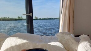 A hand holding a mug, a bed with white bedding, and a view of a body of water through a window. - Fantastic Houseboat `Splendid` with Tender Kaag (Monnickendam)