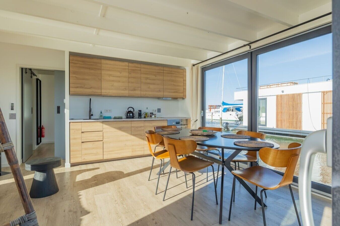 Una cocina moderna con armarios de madera, una zona de comedor con mesas y sillas redondas, y vistas a barcos a través de amplios ventanales.