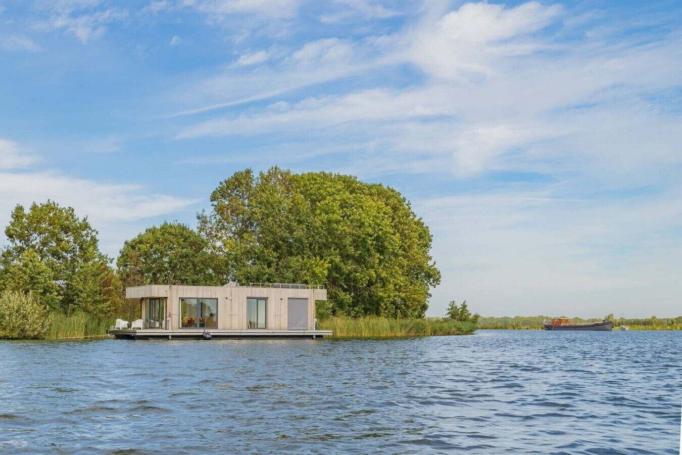 Une maison flottante au design moderne, située sur une étendue d’eau calme, entourée d’une végétation luxuriante et sous un ciel dégagé.