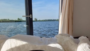 A hand holding a mug on a bed with a view of a body of water through a window.