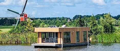 A floating houseboat with a deck and windows, situated on a body of water with a windmill in the background.