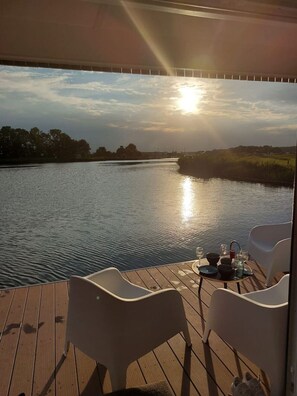 En terrass med två stolar och ett bord dukat för middag, med utsikt över en stilla flod och en solnedgång i bakgrunden.
