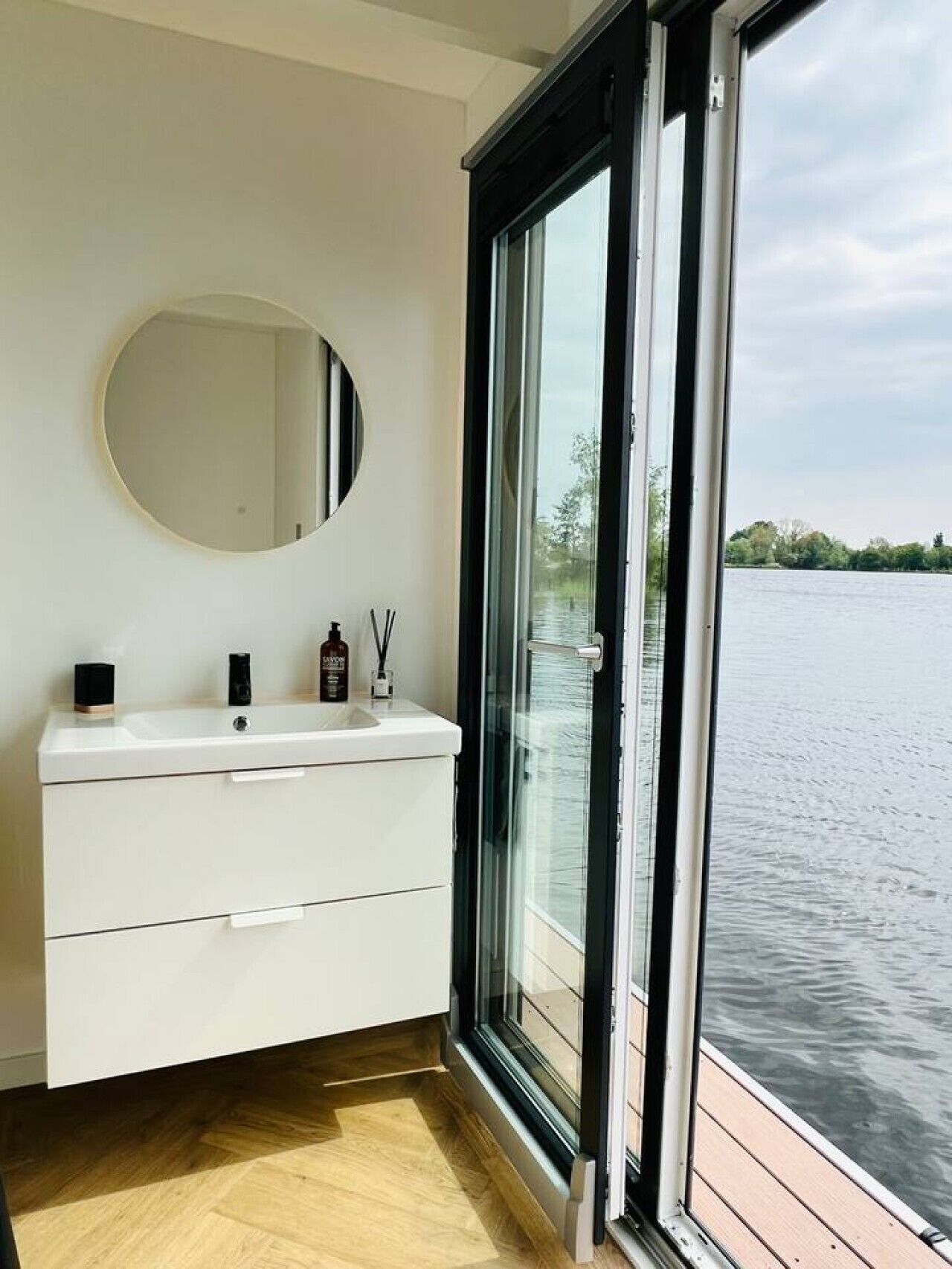 Ein Badezimmer mit weißem Waschtisch, einem runden Spiegel und einem großen Fenster mit Blick auf ein Gewässer.