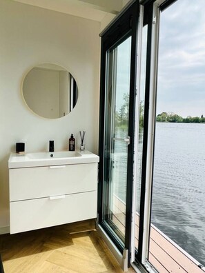 A bathroom with a white vanity, a round mirror, and a large window offering a view of a body of water.