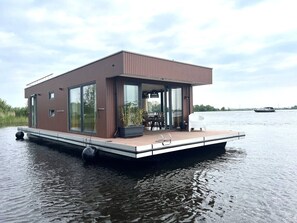 Ett flytande hus med en terrass, stora fönster och en vattenyta i bakgrunden.
