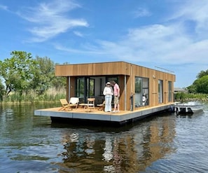 Une maison flottante dotée d’une terrasse, de chaises longues et d’un petit bateau amarré à proximité.