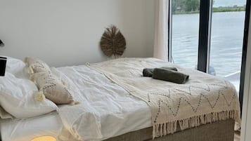 Een slaapkamer met een bed, een bedlampje, uitzicht op een waterpartij door een raam en een klein krukje.