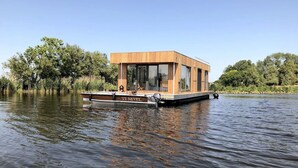 A houseboat with a wooden exterior, large windows, and a flat roof, floating on calm water.