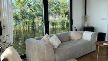 Een moderne woonkamer met een bank, twee schommelstoelen en uitzicht op een waterpartij door grote ramen.