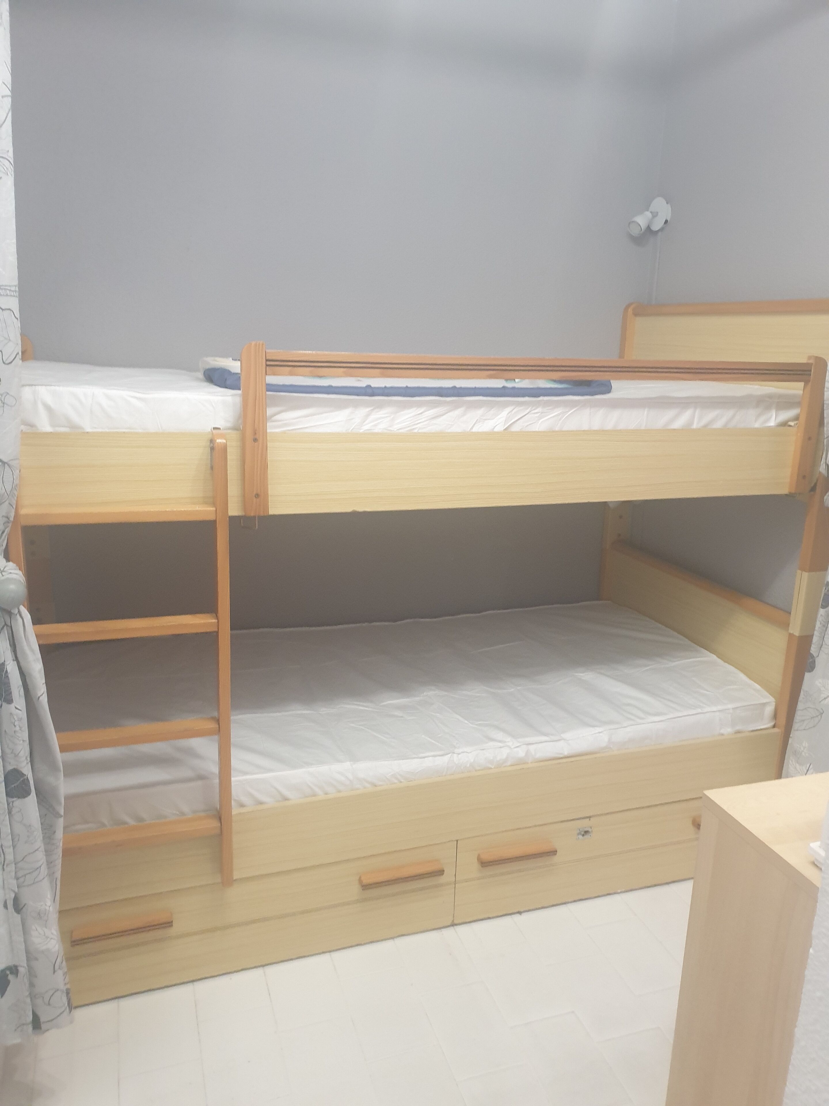 A bunk bed with a ladder and drawers, a curtain, and a small white object on the wall.