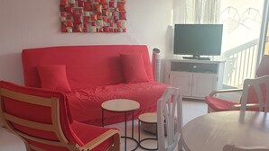 Una sala de estar con un sofá rojo, un sillón de madera, una mesa de comedor redonda y un televisor sobre un mueble.