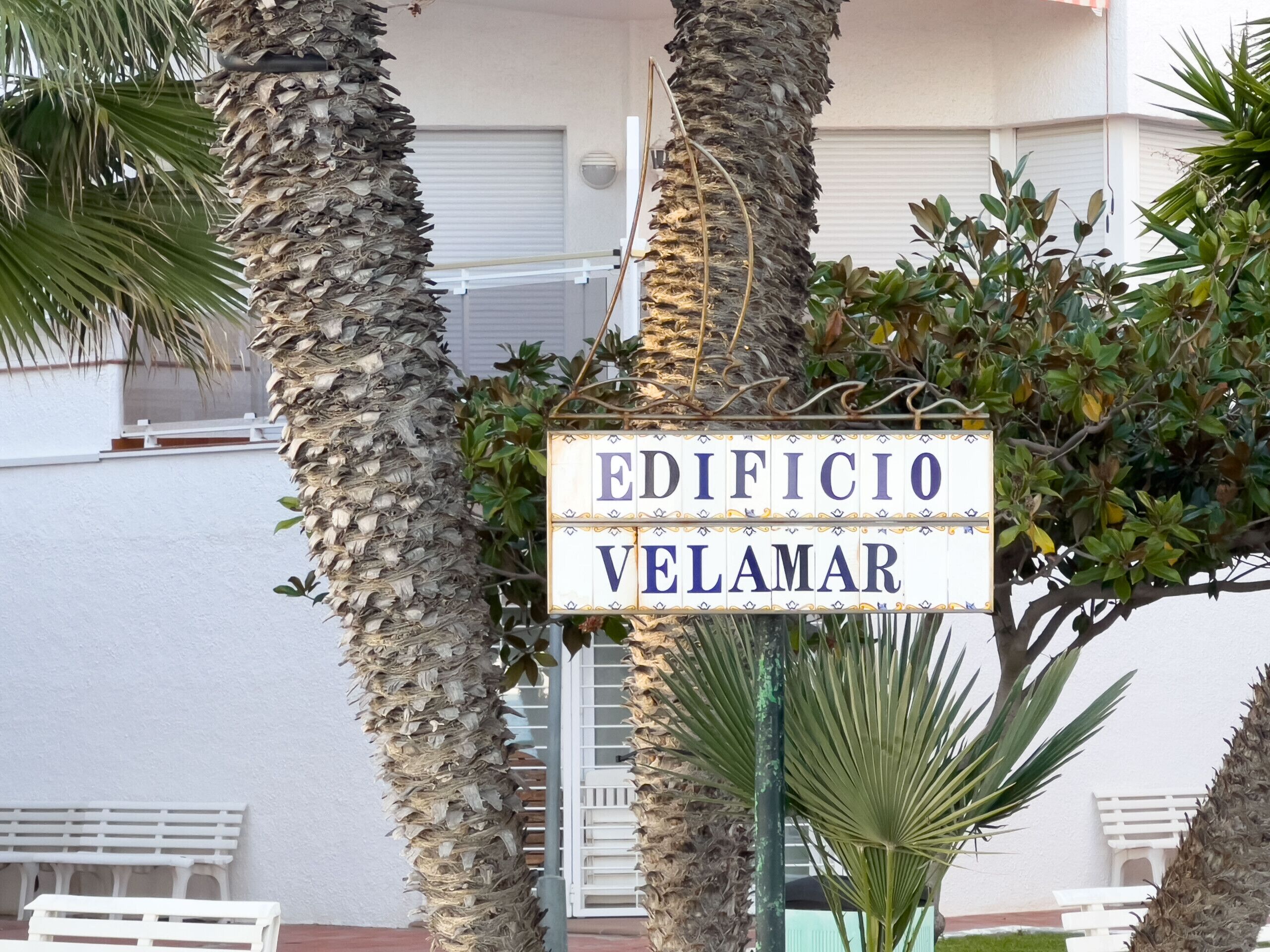 Un cartel que dice 'EDIFICIO VELAMAR', rodeado de palmeras y un edificio al fondo.