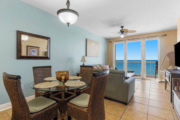 A dining area with wicker chairs and a glass table, a living room with a sofa and a view of the ocean, and a balcony with a view of the sea.