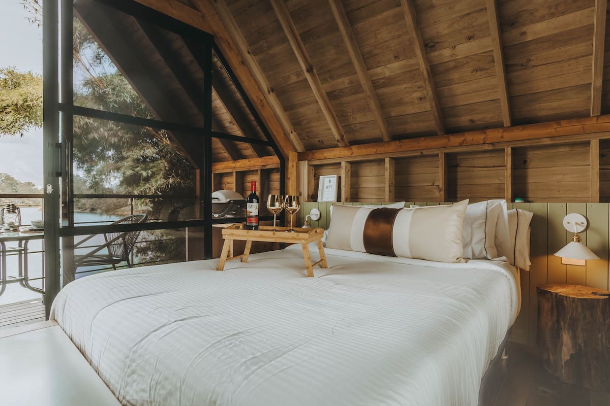 A bedroom with a large bed, wooden beams, a wooden nightstand, and a view of the outdoors.