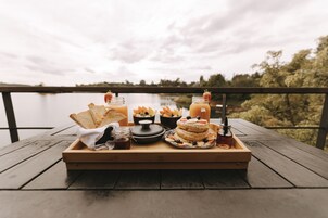 A wooden tray with food and drinks on a wooden deck by a lake.
