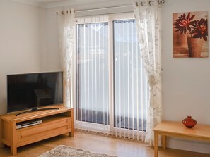 A living room with a TV on a wooden stand, a sliding glass door with white curtains, a small table with a vase, and a painting on the wall.
