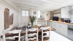 On aperçoit un coin repas avec une table en bois dressée pour quatre personnes, entourée de chaises blanches. Au fond, on distingue une cuisine avec des meubles de couleur claire et un mur d’accent en pierre.