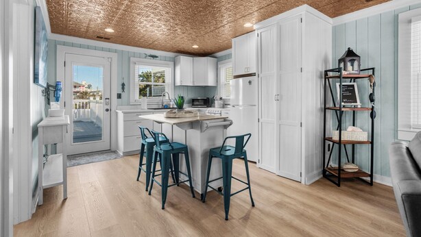 A modern kitchen with white cabinets, a central island with bar stools, a wooden ceiling, and a view of the outside. - Rocks On The Side: Private Pool, Steps to the Beach, Murrells Inlet (Murrells Inlet)