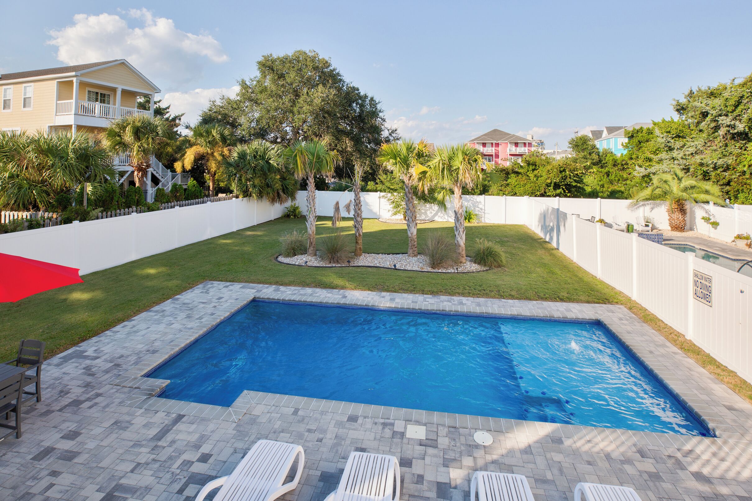 Piscina rectangular con un borde de piedra, rodeada por una cerca blanca y palmeras.