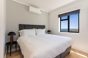 A bedroom with a large bed, two bedside lamps, a window with a view, and an air conditioning unit.