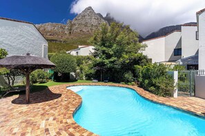 A swimming pool with a brick deck, surrounded by white buildings, trees, and a mountain in the background.