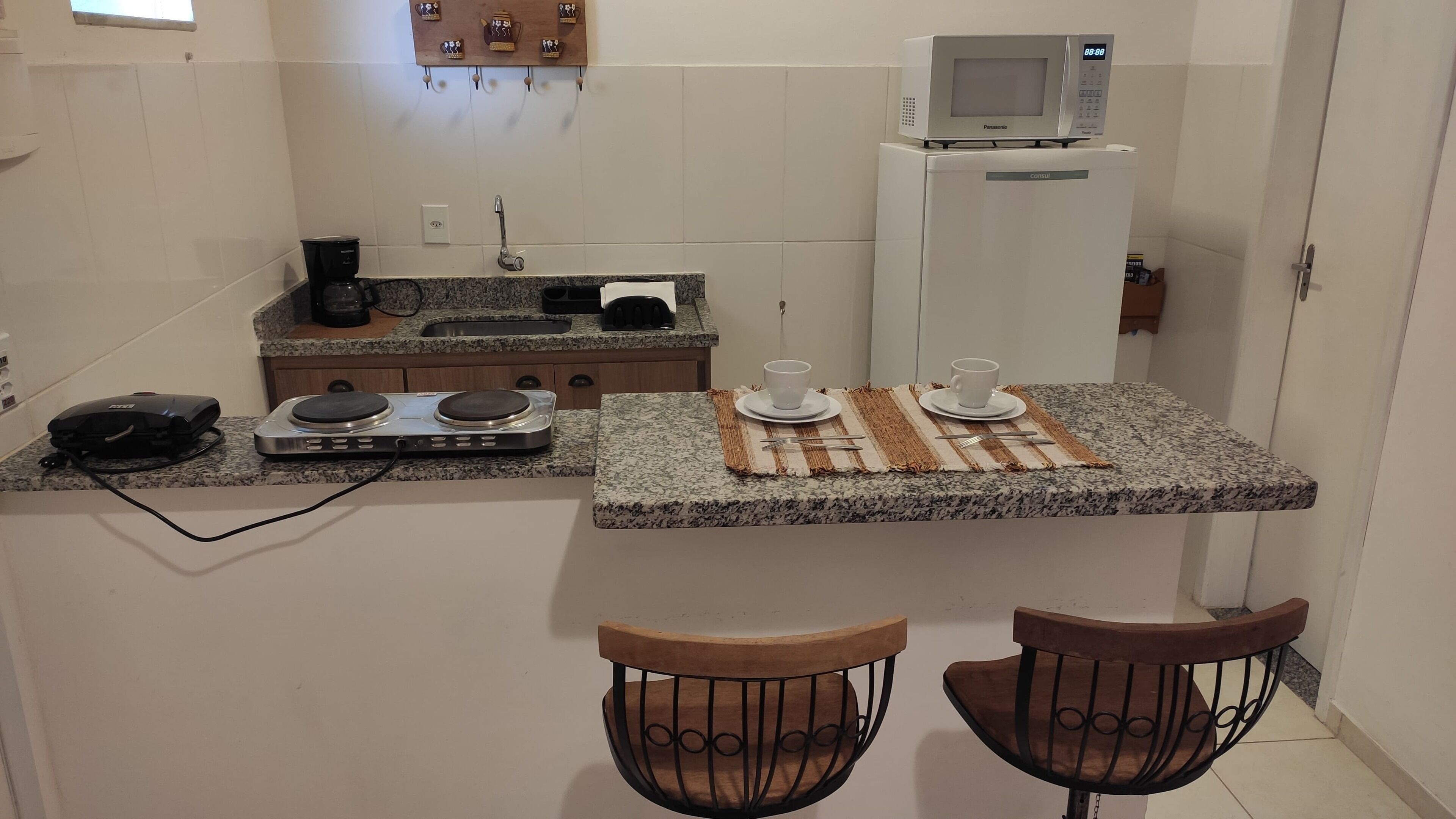 Une cuisine compacte équipée d’un four à micro-ondes, d’une machine à café et d’un petit réfrigérateur. On y trouve deux tabourets et une petite table avec des tasses et des couverts.