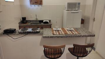Une cuisine compacte équipée d’un four à micro-ondes, d’une machine à café et d’un petit réfrigérateur. On y trouve deux tabourets et une petite table avec des tasses et des couverts.
