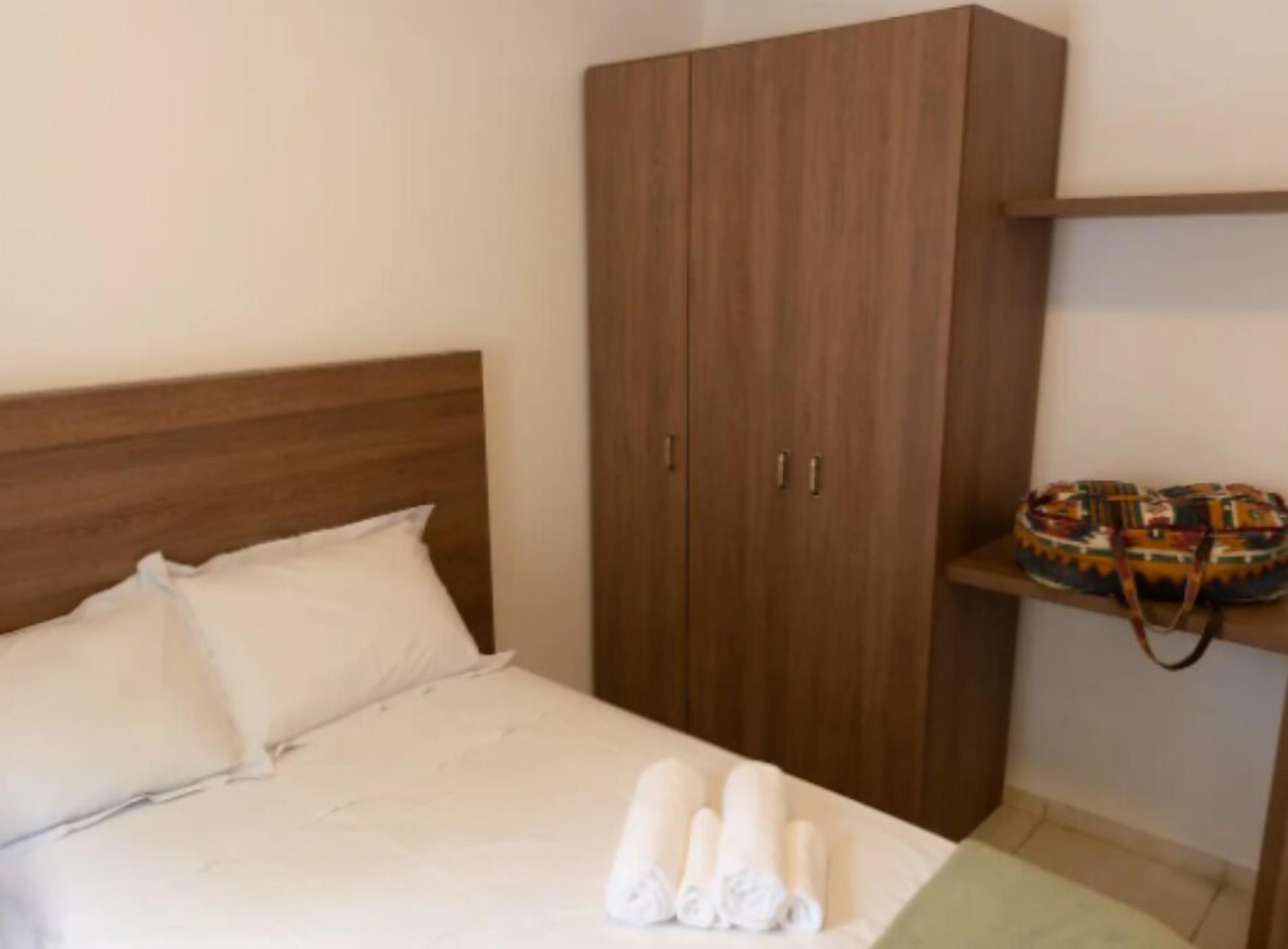 A bedroom with a wooden headboard, a bed with white linens, a wardrobe with two doors, and a shelf with a patterned bag.
