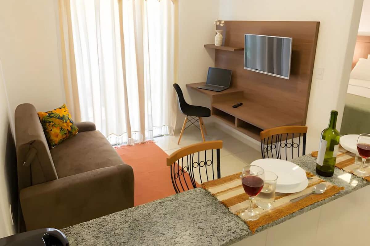 A modern living room with a sofa, a dining table with chairs, a TV mounted on the wall, and a kitchen area with a wine bottle and glasses.