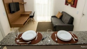 Un espacio habitable compacto con kitchenette, un sofá, un televisor montado en un estante de madera y una zona de comedor con dos platos y vasos.