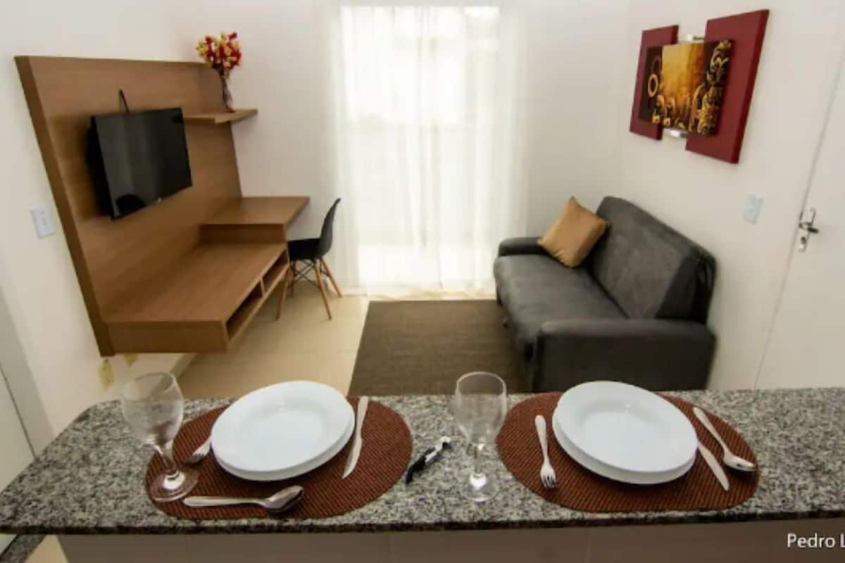 A compact living space with a kitchenette, a sofa, a TV mounted on a wooden shelf, and a dining area with two plates and glasses.