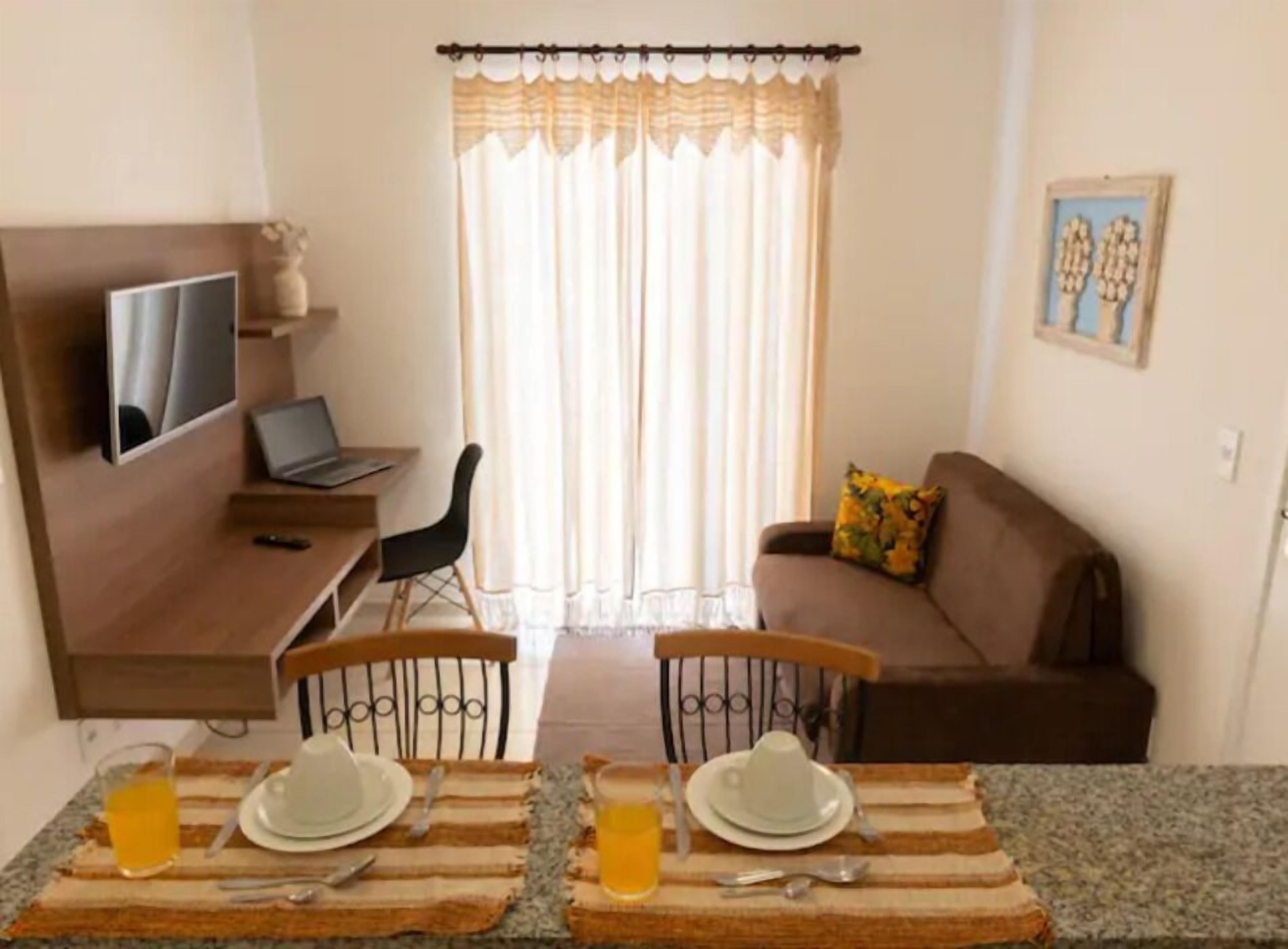 A small, cozy living area with a sofa, a TV mounted on the wall, a dining table set with plates and cutlery, and a window with curtains.