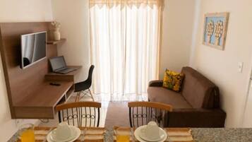 A small, cozy living area with a sofa, a TV mounted on the wall, a dining table set with plates and cutlery, and a window with curtains.