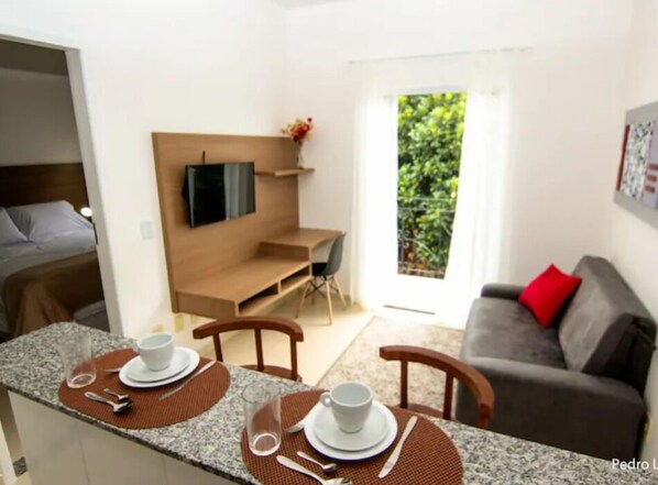 A modern living room with a dining area, a sofa, a TV mounted on the wall, and a view of greenery outside. - Apart Sol Nascente (Itatiaia)