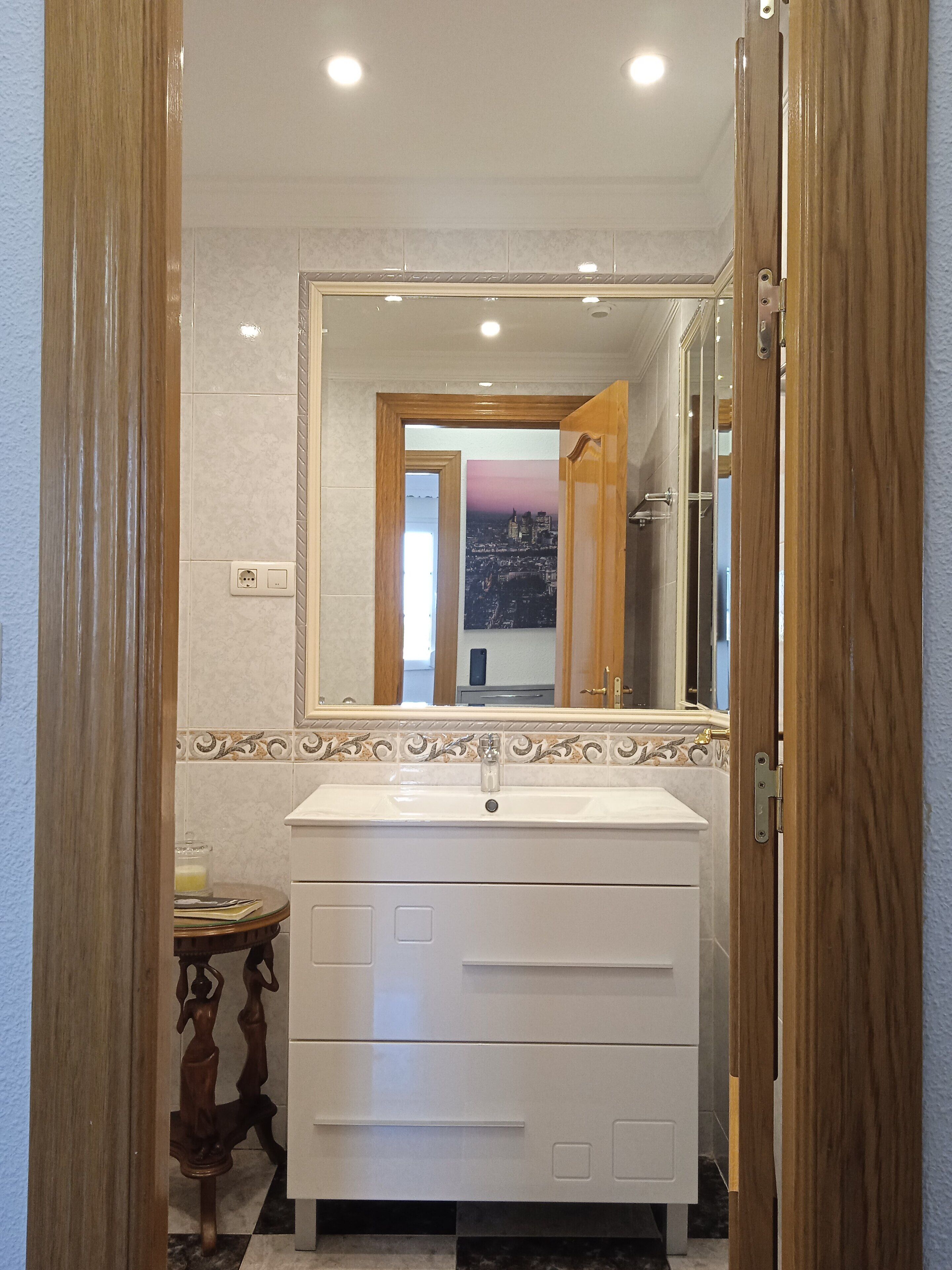 Casa de banho com lavatório branco, porta de madeira e espelho refletindo uma vista urbana.