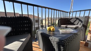 Un balcó amb mobles de vimet, una taula amb pomes i una revista, i una vista despejada dels edificis i turons circumdants.